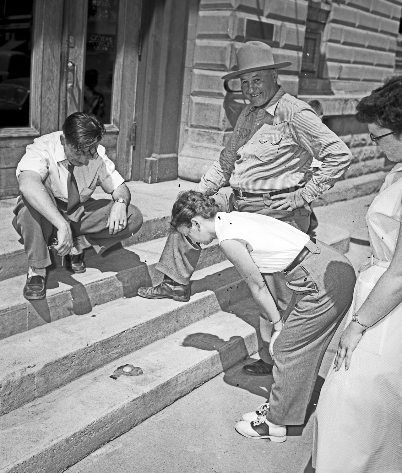 A black and white photo shows several people in dated clothes looking at an egg that has been cracked on the steps of a sandstone building. A chubby man in a hat looks into the camera with a large smile.