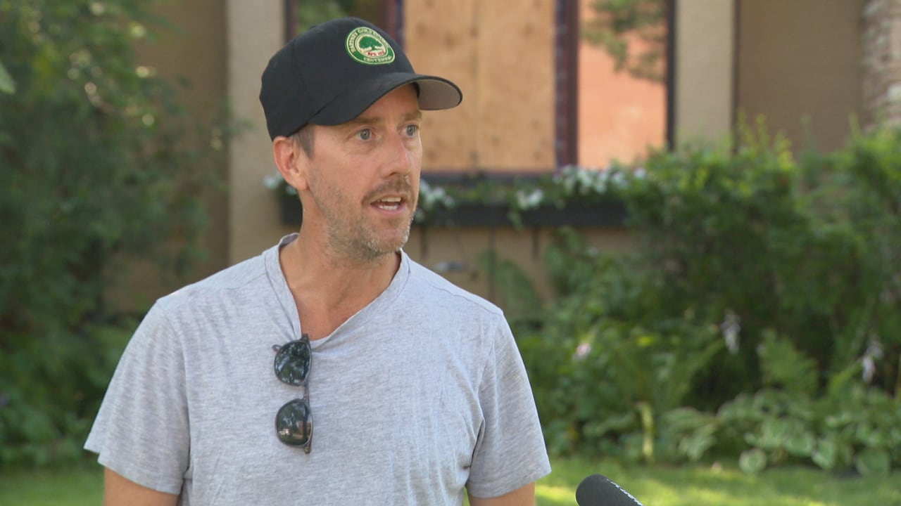 A man in a T-shirt and a baseball cap speaks outside a house with boarded-up windows.