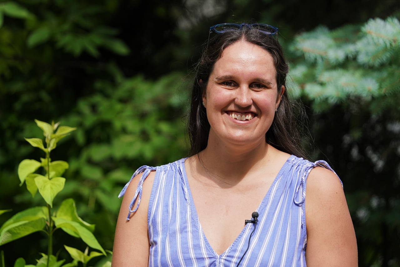 A woman with long brown hair smiles standing next to trees and shrubs.