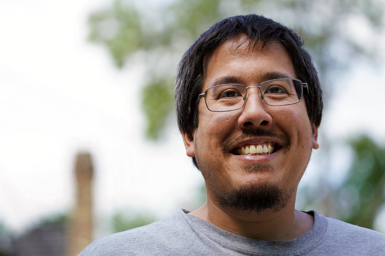 A man with glasses and facial hair is seen smiling outdoors.
