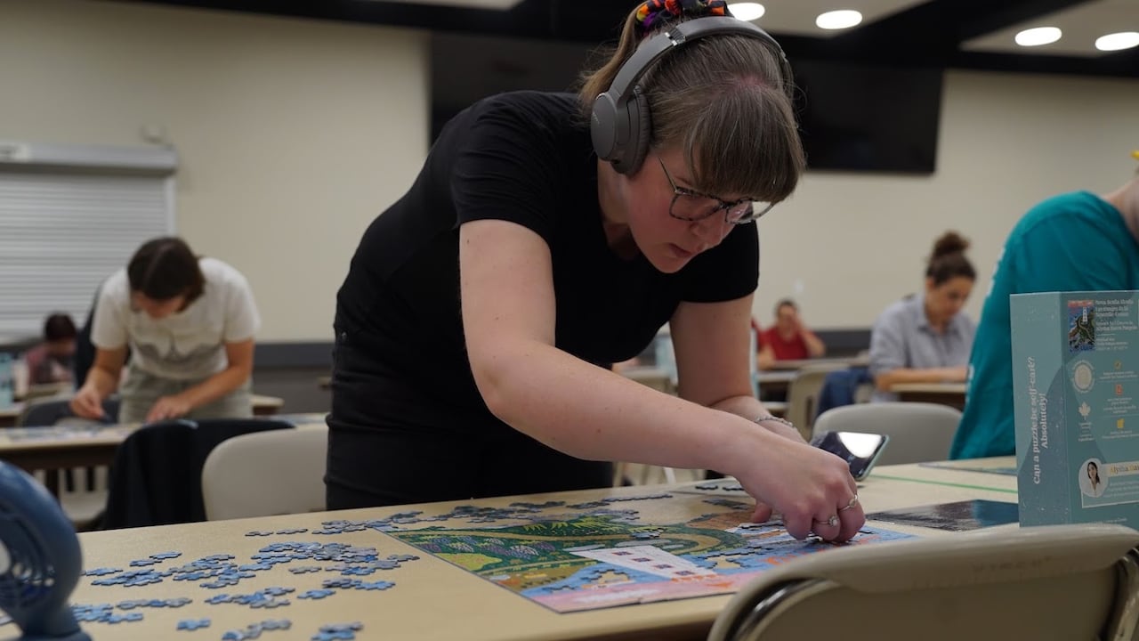 A woman wearing headphones and glasses stands in front of a table while putting a jigsaw puzzle together.