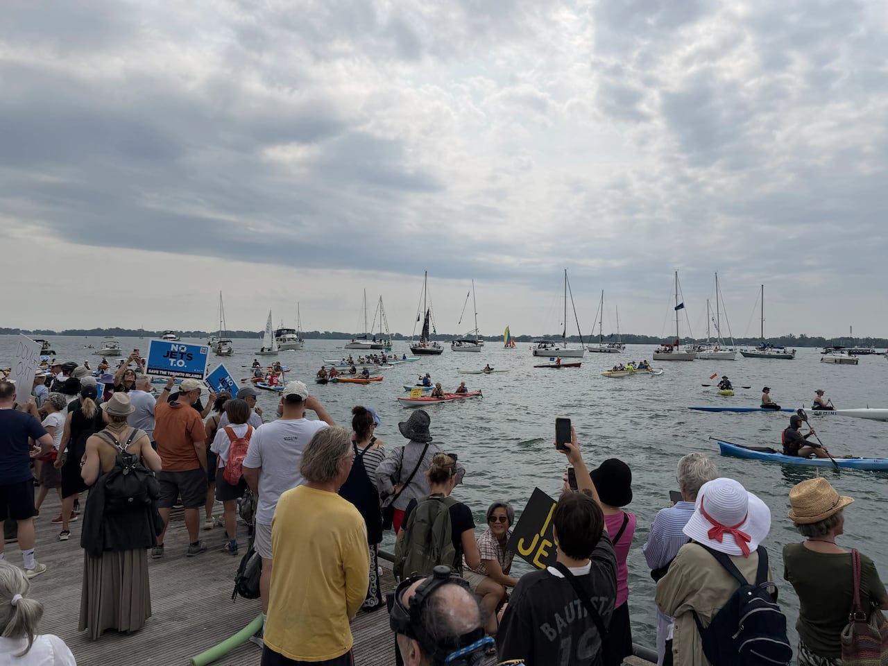 People standing by pier, with boaters and kayakers nearby on the water