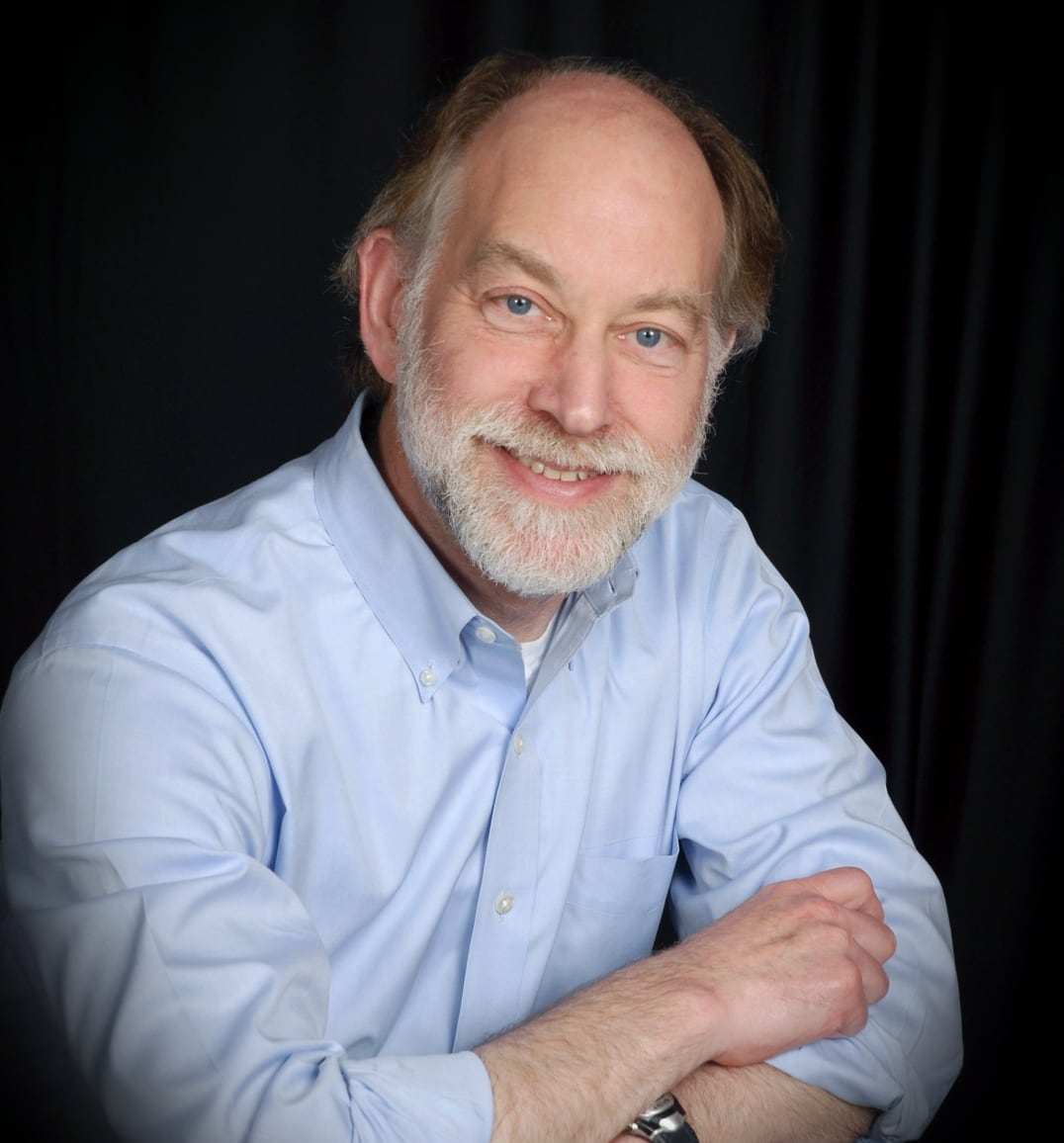 A portrait of a smiling older man with a white beard in a light blue button down shirt.