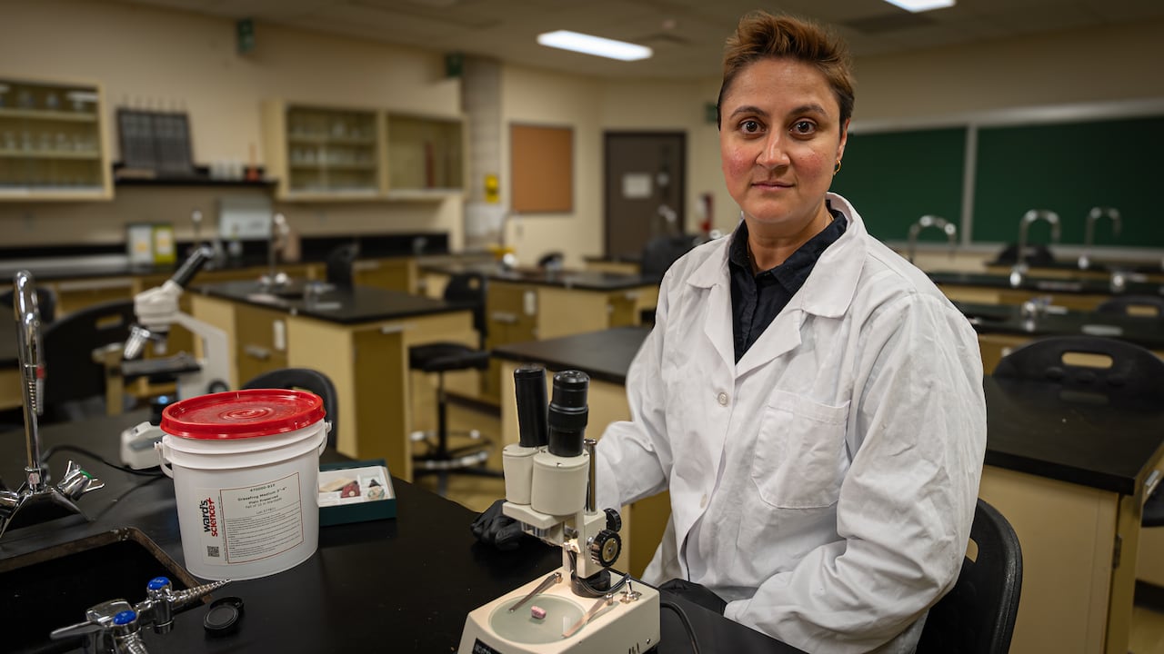 A woman with short hair, wearing a white lab coat and black protective gloves, sits in a science classroom in front of a microscope.