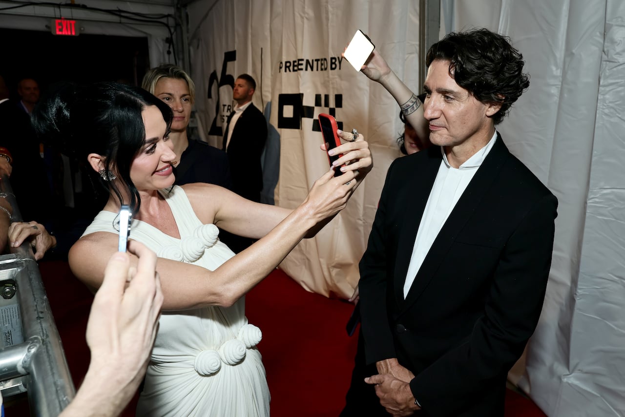 A woman in a white dress uses her cellphone to take a picture of a man in a black tuxedo.