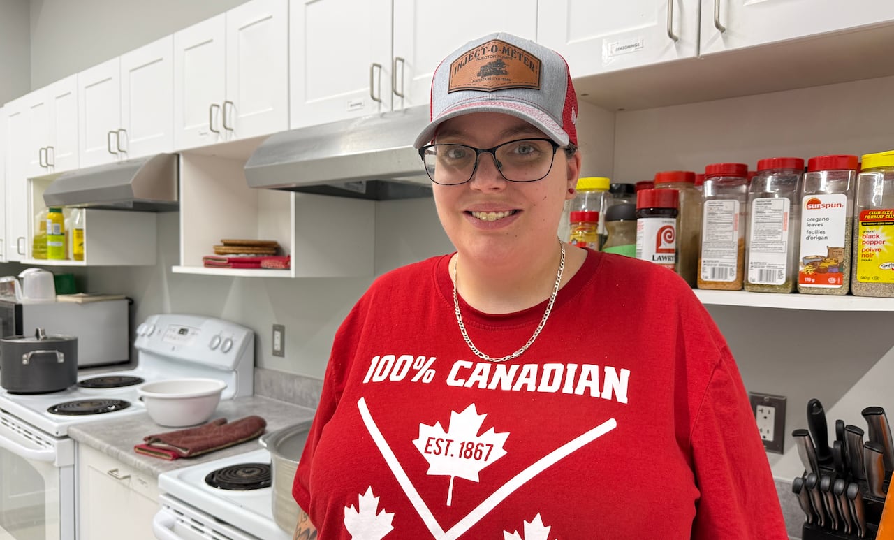 A smiling 31-year-old Caucasian woman stand in a kitchen wearing a ballcap.