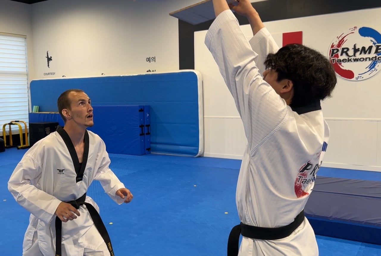 A man dressed in a white marshall arts robe, bends down and looks up to a board he's hoping to break.