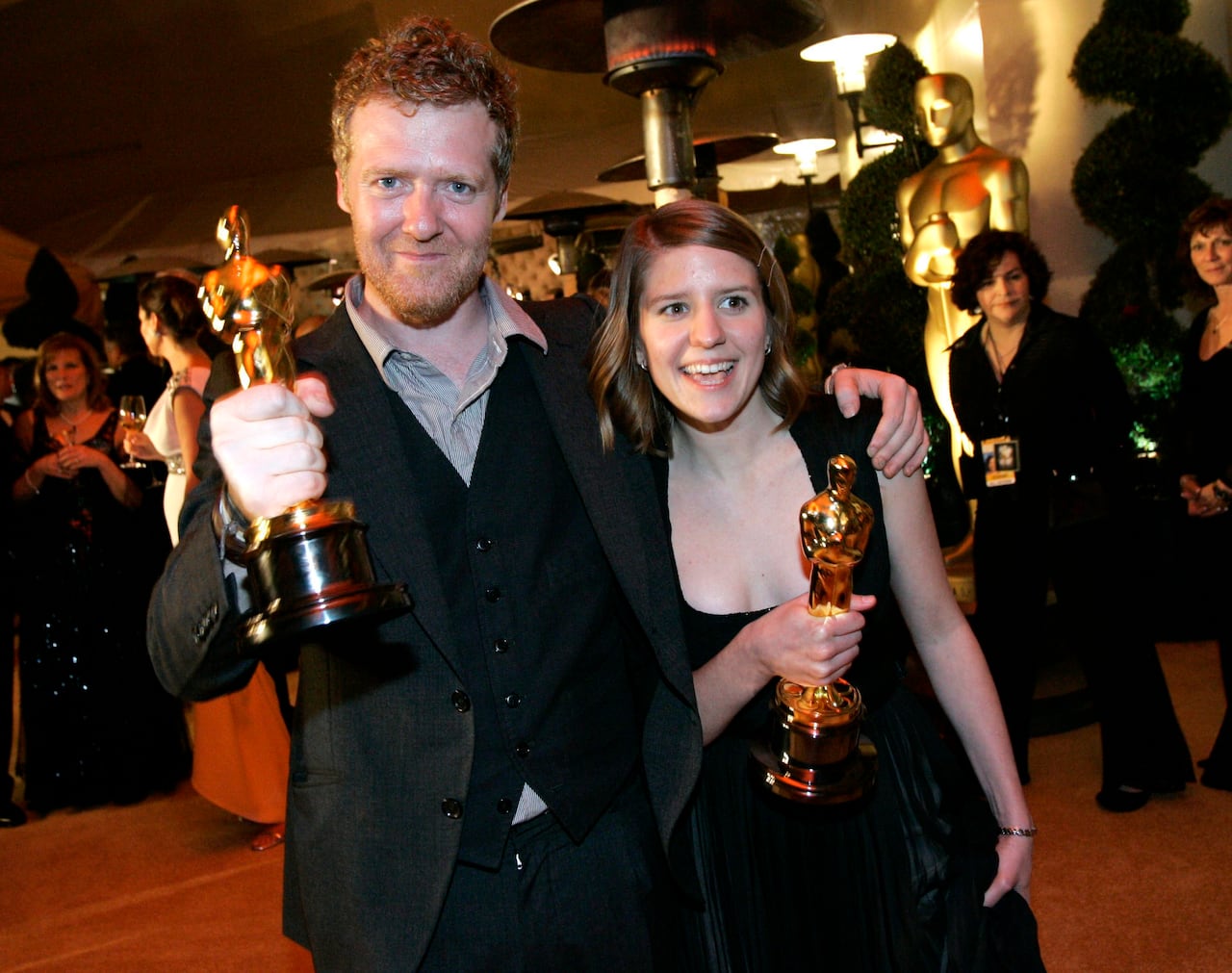 A bearded man and a dark haired woman dressed in formal wear hold up trophies.