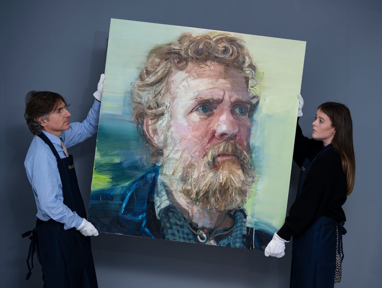 A man and a woman wearing gloves hold up a painting of a bearded Caucasian man.