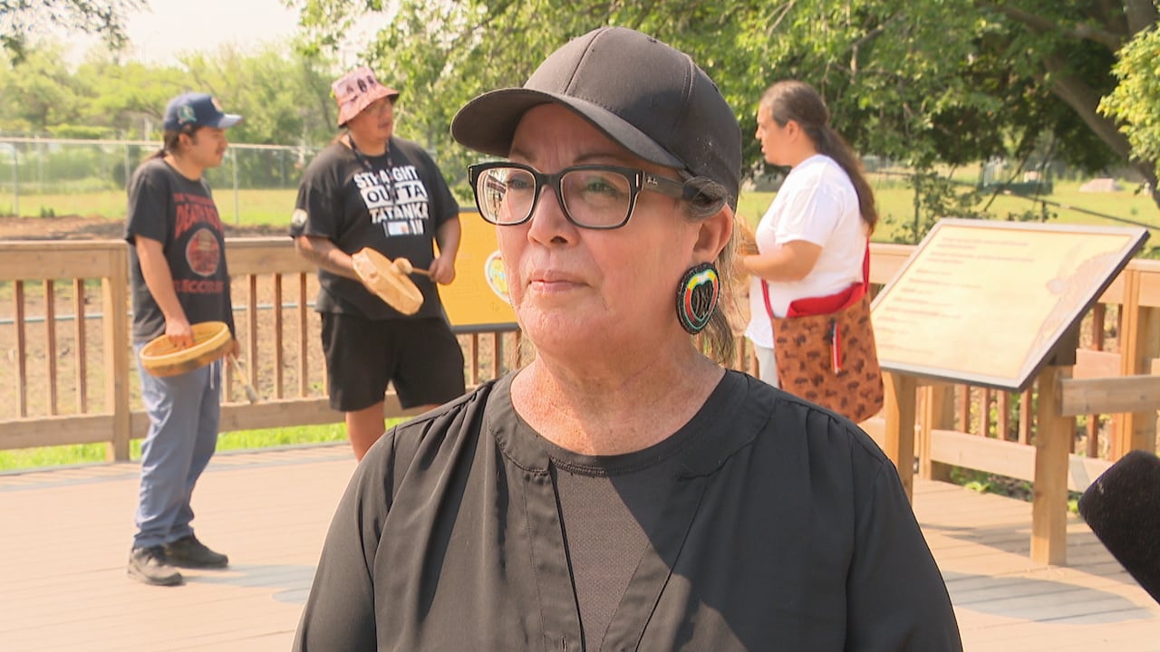 A woman wears a black t-shirt, black baseball cap and black, white, yellow and red beaded earrings while standing in front of a zoo enclosure.