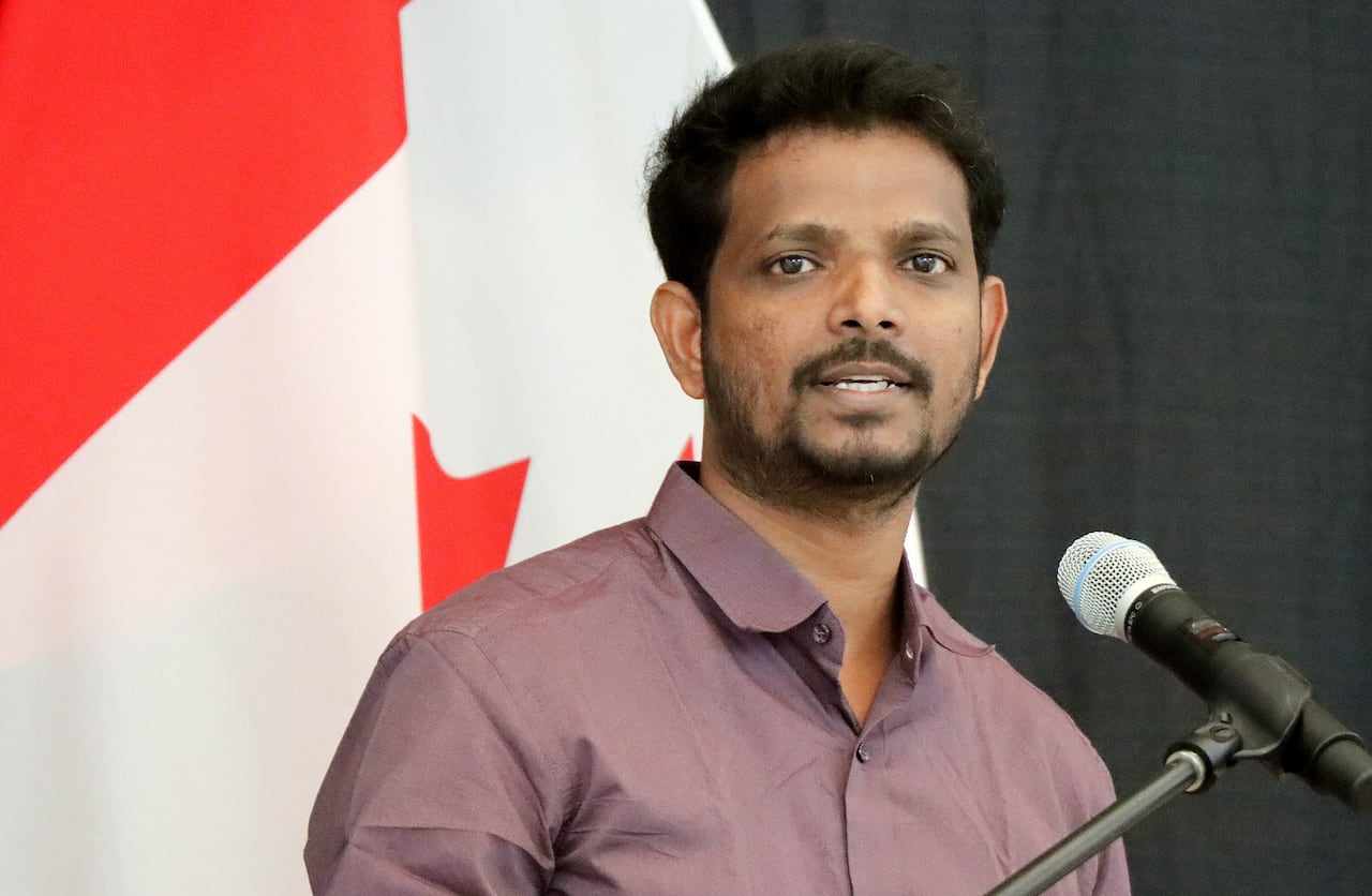 A young man with black hair, beard and moustache wearing a maroon shirt speaks into a microphone i front of a red and white Canadian flag.