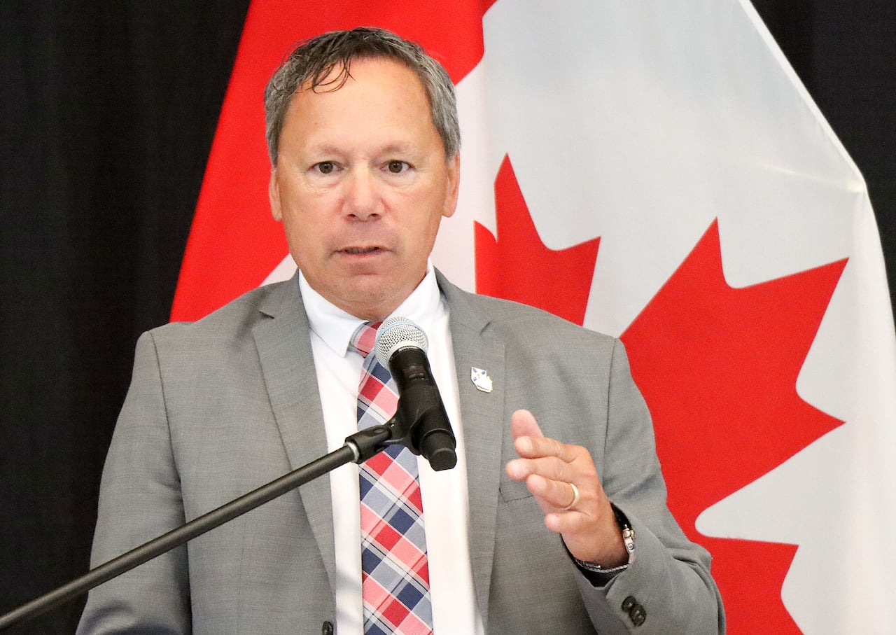 A man in a grey suit jacket with a white shirt and plaid tie gestures and speaks at a microphone in front of a red and white Canadian flag.