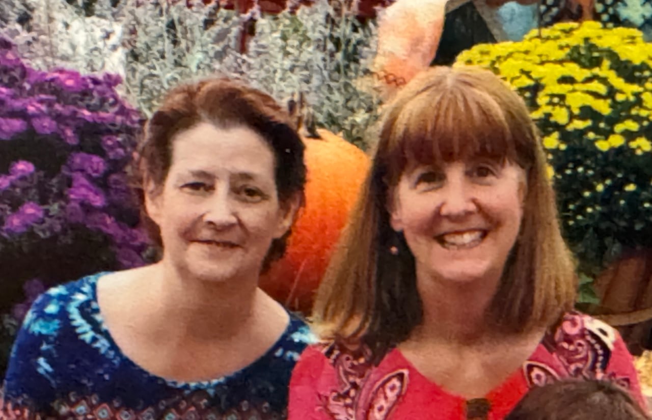 Two smiling, middle-aged woman, one with short red hair and one with a long brown bob, smiling in front of flowers and a pumpkin.