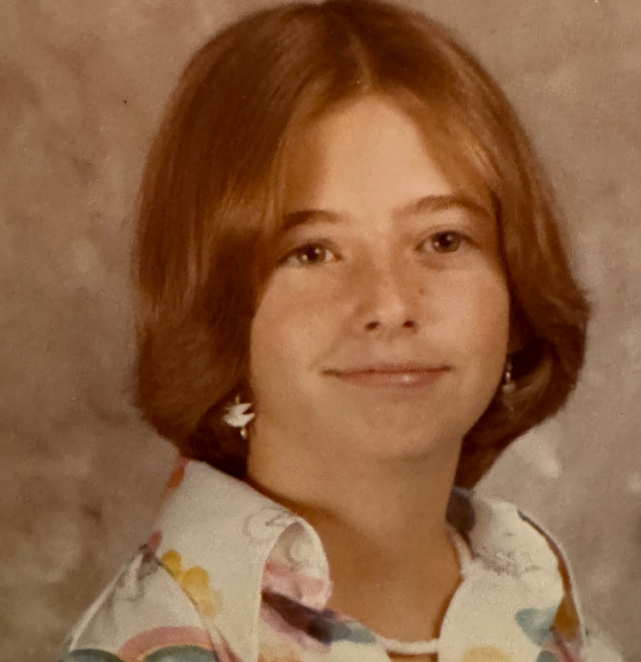 An 1970s school photo of a young girl with red hair in a floral print blouse.
