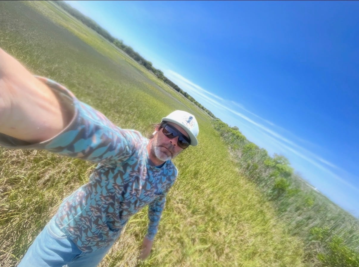 A man with a white beard, dark sunglasses and a ballcap takes a selfie in a big open field.