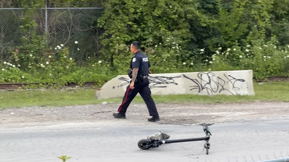 A Toronto police officer walks by a toppled e-scooter on an empty city street. There are only trees and a cement barricade in the background. It is evening time in the summer.