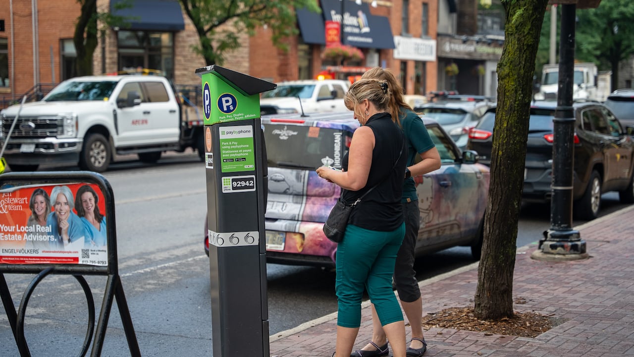 Two people pull out a wallet in front of a parking metre in a city on a cloudy summer day.