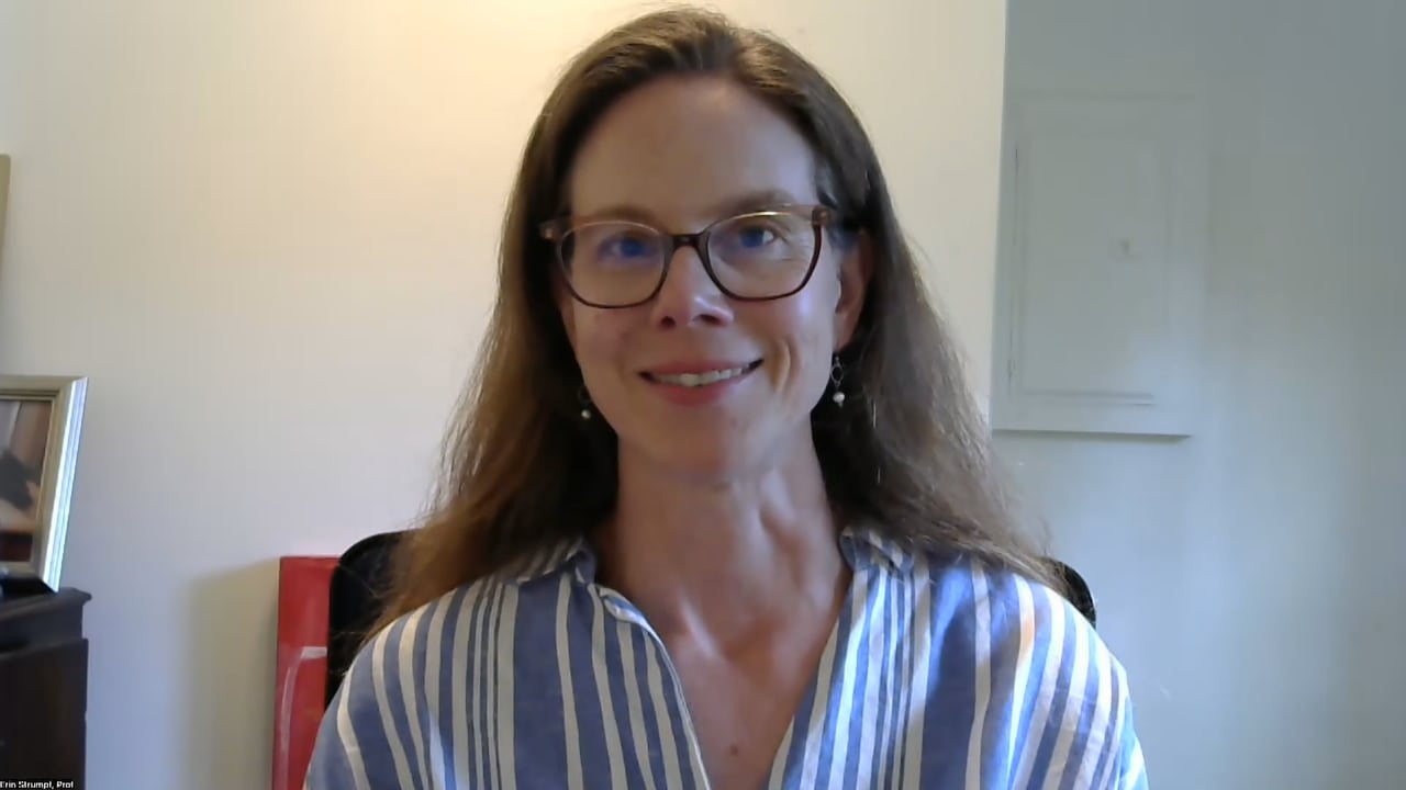 A woman with brown hair and light brown glasses sits smiling at the camera. She is wearing a blue striped shirt and dangling earrings.
