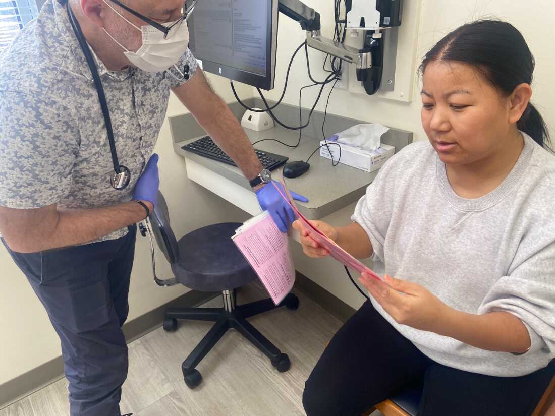 Pediatrician John Snyder stands on the left side of the frame, while Tenzin Dekey, who is seated on the right side of the frame, holds papers with vaccine information.