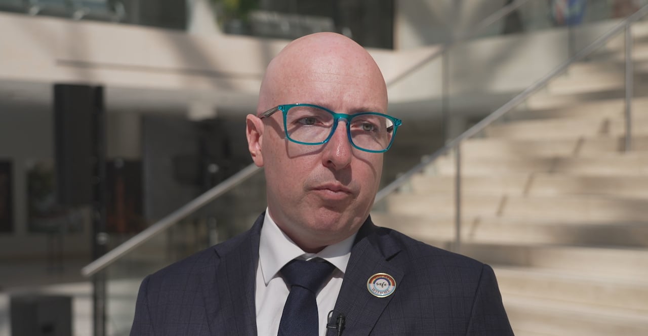 A white man wearing a navy suit looks at a camera. He wears blue glasses and is bald.