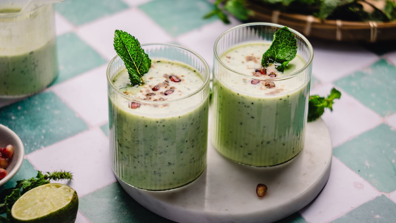 2 glasses of light green lassi on a marble board on top of a green checked tabletop. 