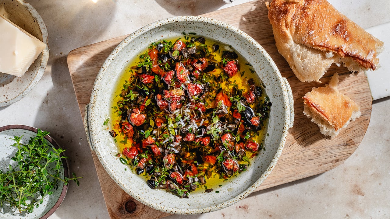 A serving bowl filled with a mediterranean dipping oil, on a wooden platter, next to a crusty loaf of torn bread.