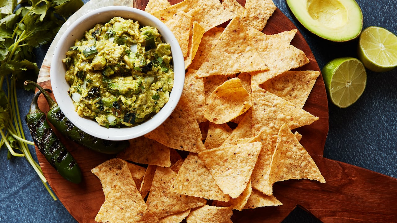 A bowl of guacamole on a wooden cutting board with tortilla chips on it. Grilled jalapeños, sliced lime and avocado sit next to it. 