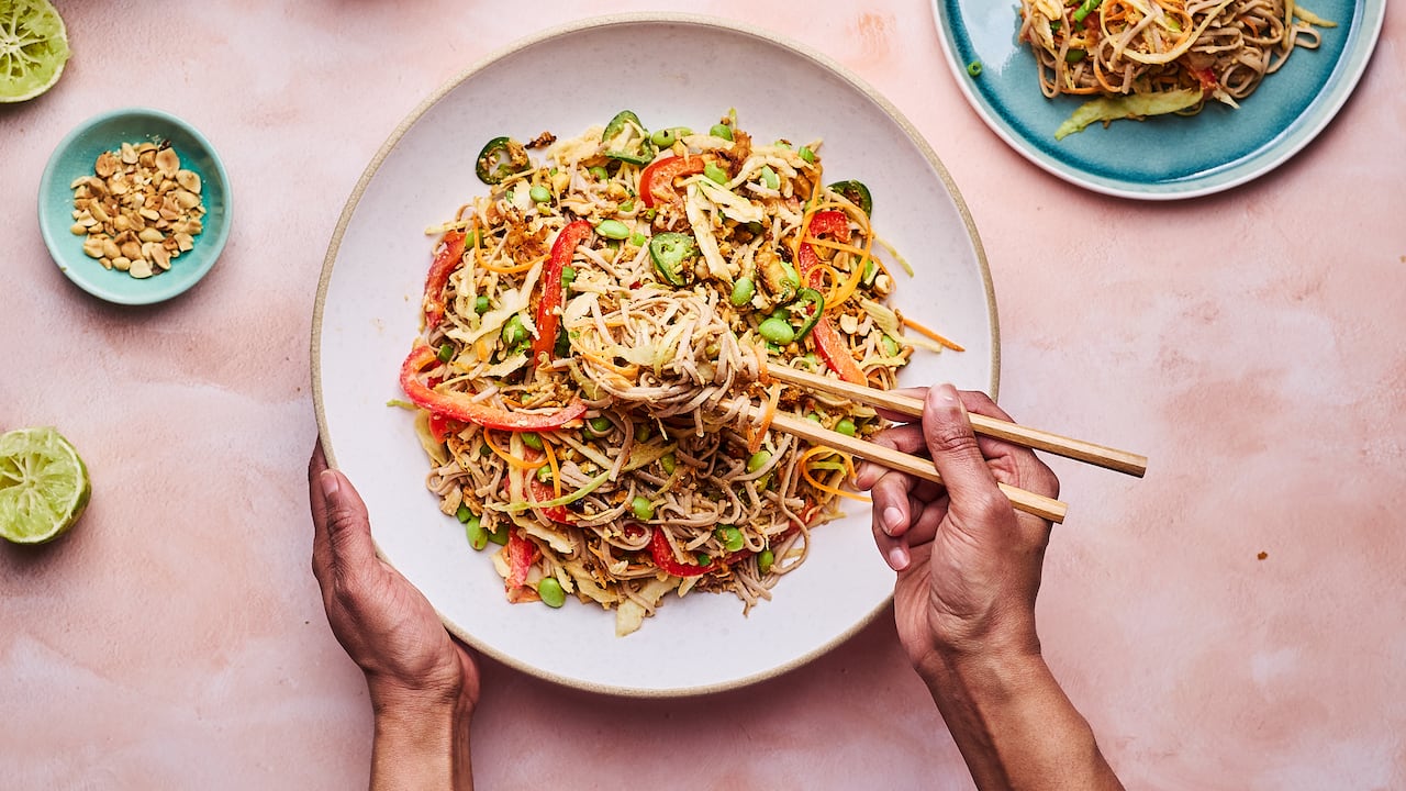 Top down shot of a bowl of soba noodle salad with slices of pepper and edamame in it. 2 hands are visible: the left hand holds the bowl and the right hand picks up noodles with chopsticks.