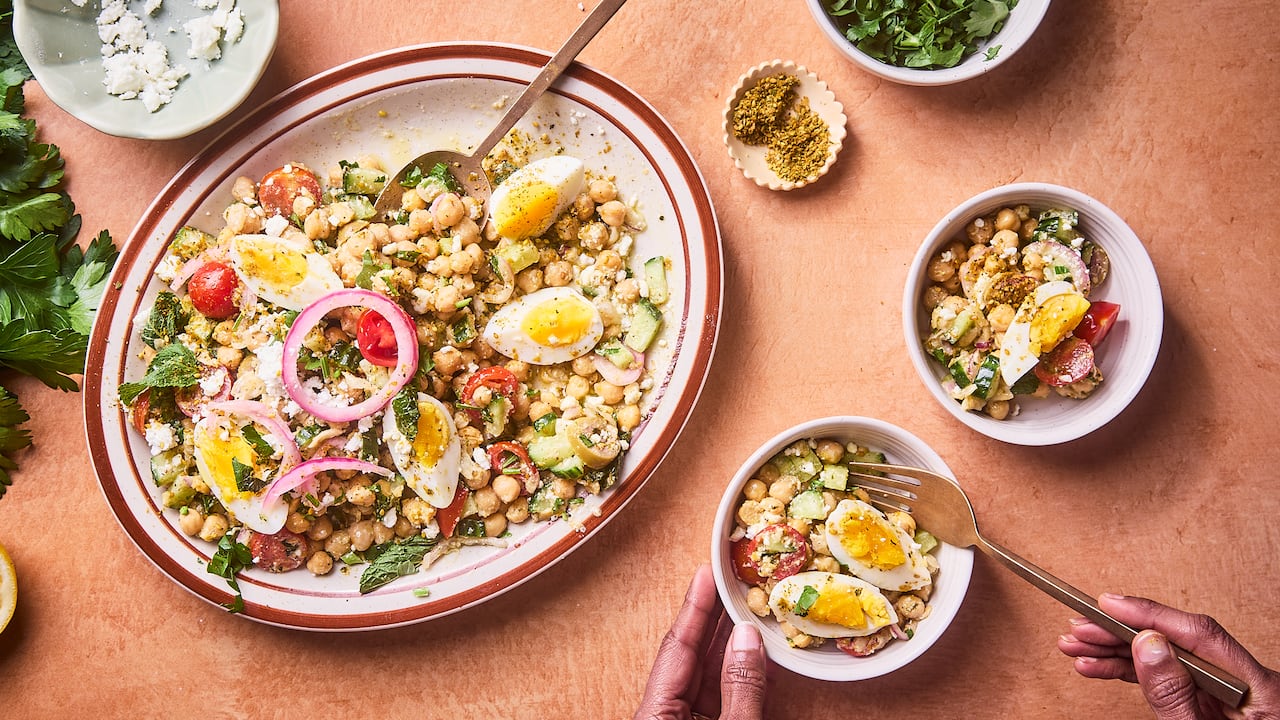 A platter of chickpea feta salad with boiled eggs and herbs on a countertop. Small bowls of ingredients and of the salad sit next to it.
