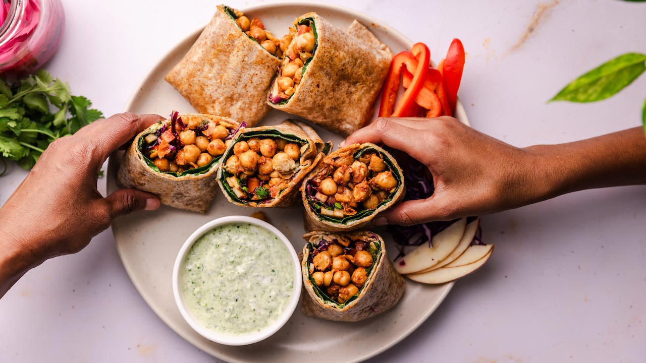 Overhead shot of a plate of chickpea wraps cut in half. 2 hands are reaching into frame to grab a wrap.