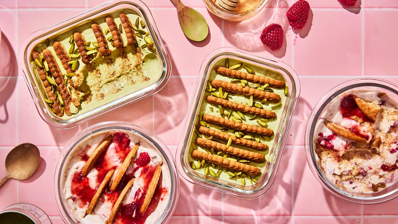 Top down shot of 4 glass containers of no-bake cheesecake on a pink tile surface. 2 have cookies and pistachios. 2 have cookies and jam.