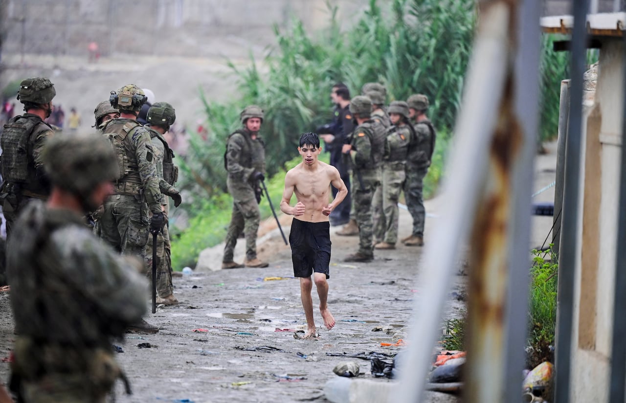 A young man in black shorts and no shift runs barefoot along a path, which is lined with armed soldiers.