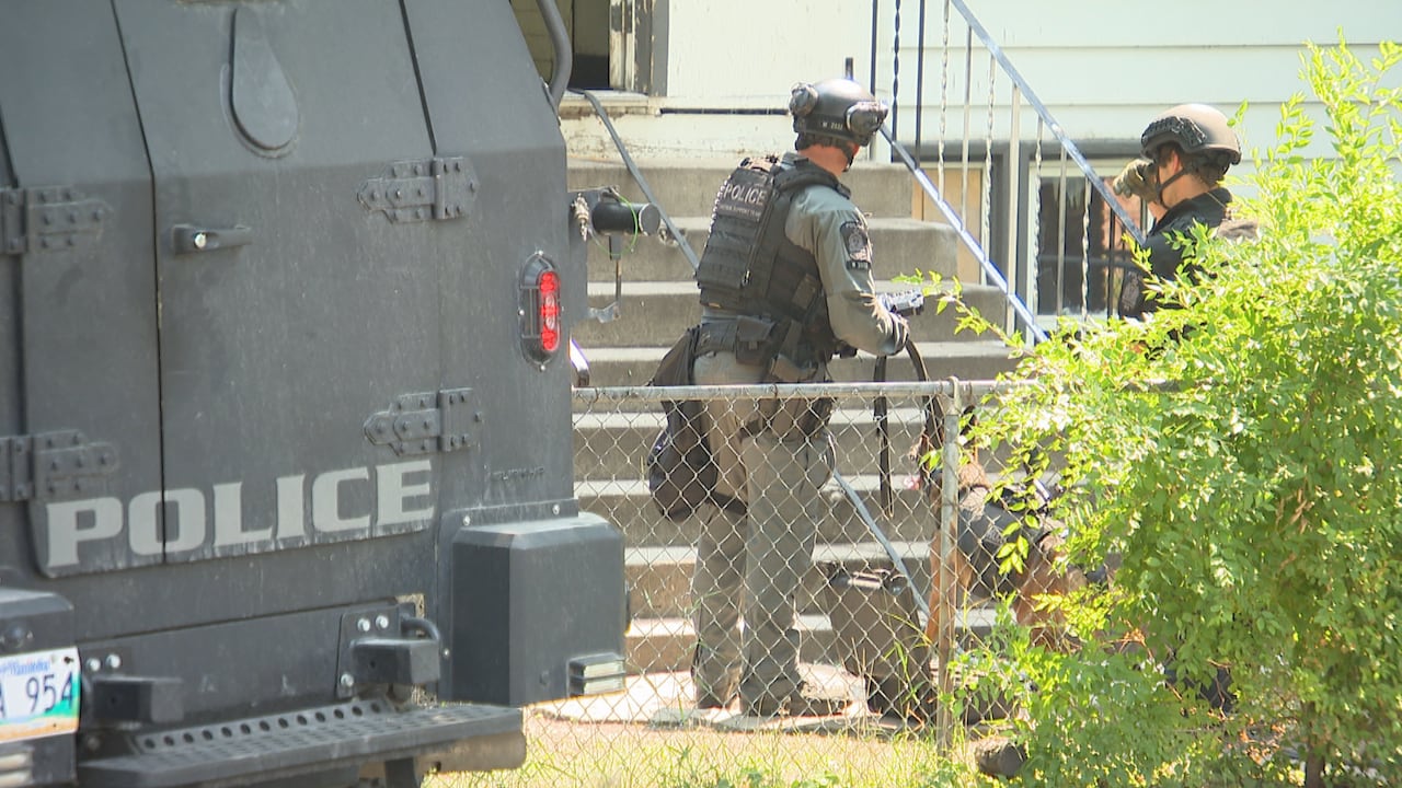 Three police officers stand in the front yard of a home, with a police dog and armoured vehicle.