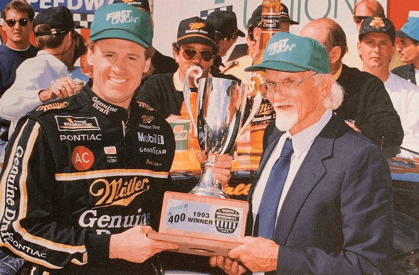 NASCAR driver in a black Miller Genuine Draft suit poses with a large trophy as an older man in a suit presents it at a victory celebration, 1993 First Union win plaque visible on the base.