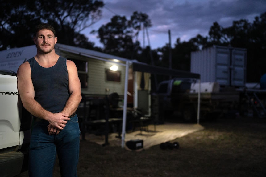 Luke Bateman wears jeans and navy singlet standing outside a caravan with light on inside at night time.
