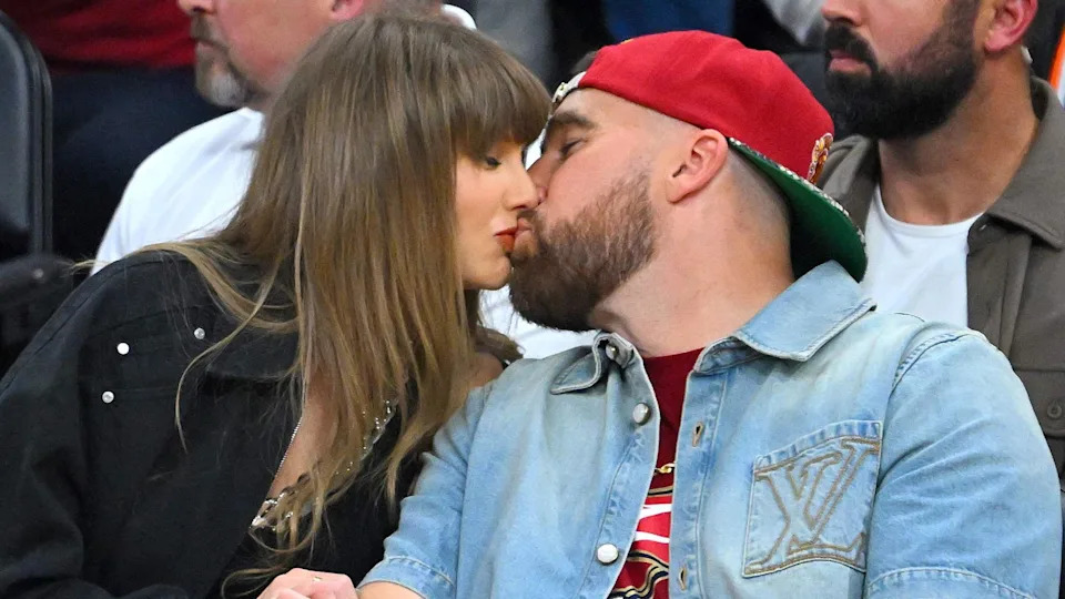 Taylor Swift and Travis Kelce kiss during a game between the New York Knicks and the Cleveland Cavaliers in Cleveland, Ohio, earlier this year. She is wearing a black jacket, he is wearing a blue deim shirt and red baseball cap.