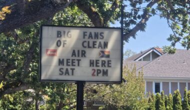 A letterboard sign reading "Big fans of clean air meet here, SAT 2 PM." IN the background is a big oak tree, blue skies and a house