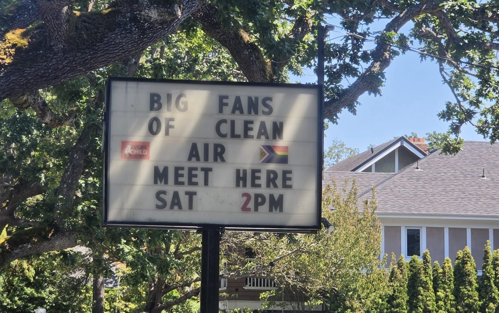 A letterboard sign reading "Big fans of clean air meet here, SAT 2 PM." IN the background is a big oak tree, blue skies and a house
