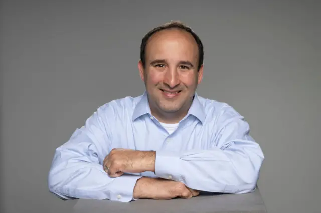 Headshot of a smiling man in a light-coloured shirt with his arms crossed in front of him
