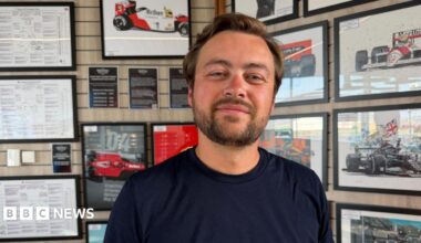 A close-up of a man wearing a navy T-shirt with the word “Takona” printed in white. He is standing indoors in front of a wall covered with framed motorsport posters, race diagrams and images of Formula 1 cars.