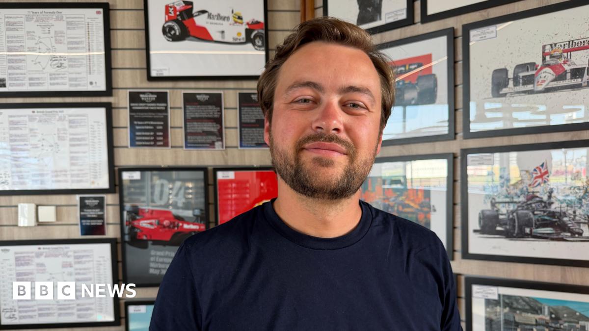 A close-up of a man wearing a navy T-shirt with the word “Takona” printed in white. He is standing indoors in front of a wall covered with framed motorsport posters, race diagrams and images of Formula 1 cars.
