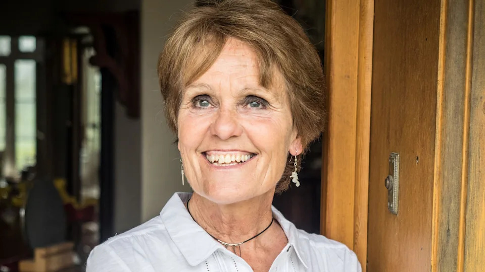 Gina Rippon. She has short brown hair and blue eyes. She is wearing a white linen shirt and black necklace, paired with seahorse shaped pendant earrings. She is leaning on a wooden door. She is smiling and looking at something out of shot.