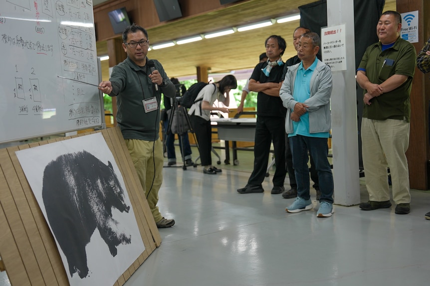 A man uses a pointing stick to mark parts of a bear displayed on a poster to a crowd of onlookers