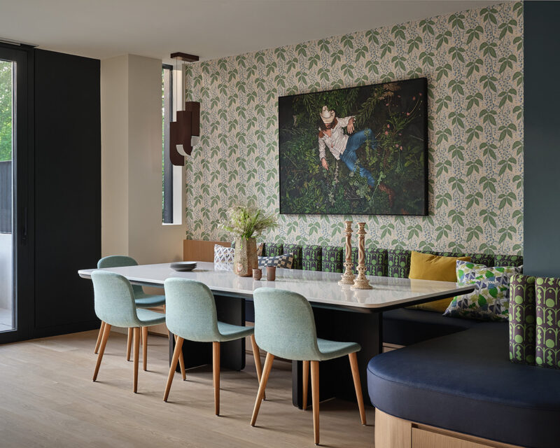 A modern dining area with a marble table, light blue chairs, patterned wallpaper, a large plant-themed photo, and built-in bench seating with assorted cushions creates an inviting space inspired by Gillian Gillies Net Zero Ready Home design.