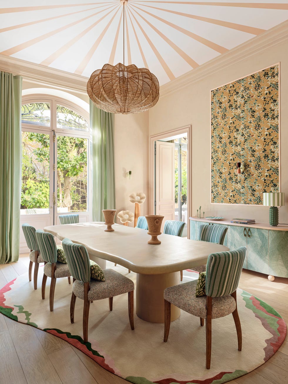 Modern dining room featuring a large table, patterned chairs, and a woven pendant light with green drapes and floral wall art.