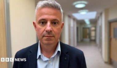 Paul Scott looks at the camera. He is standing in front of a corridor near the trust's headquarters. It is an inpatient unit. You can see a new beige corridor with light colured wood doors. Scott is wearing a navy suit jacket with a pale blue shirt