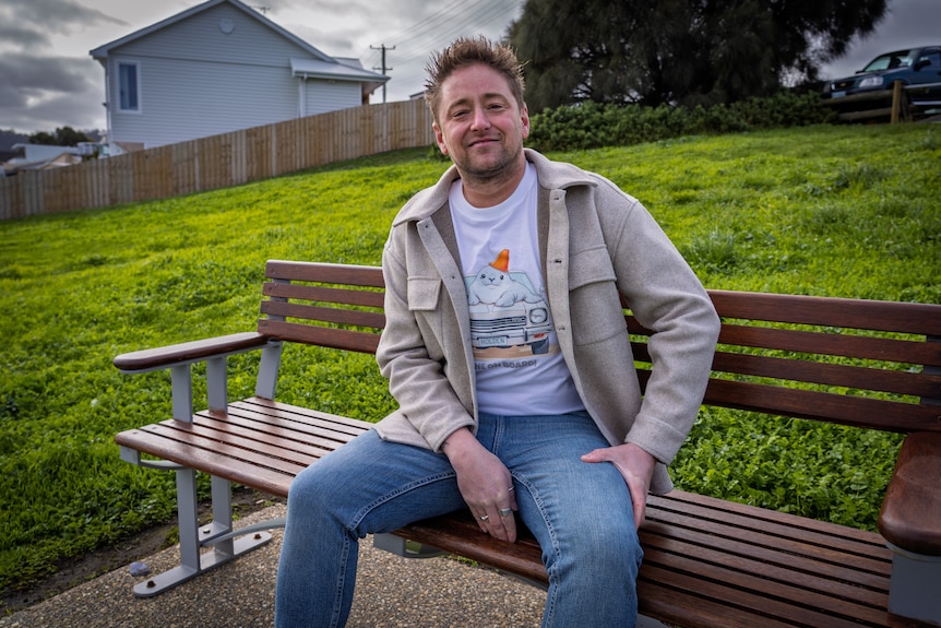Cory Young wears jeans and a T-shirt with a picture of a seal sitting on a car bonnet and sits on a park bench.