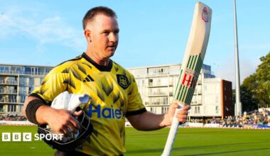Gloucestershire's D'Arcy Short walks off the pitch at the The Seat Unique Stadium, Bristol, with his bat raised in his left hand and his helmet and gloves under his right arm.