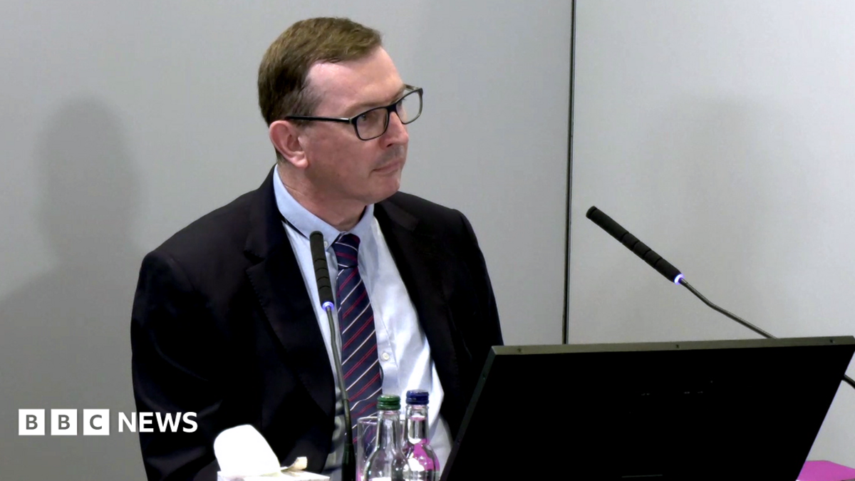 Brian O'Donnell sits behind a desk at the Lampard Inquiry at Arundel House in London. He is wearing a navy and burgundy tie and a black suit jacket, and a black laptop sits in front of him at the table. There are two bottles of water on the table and a microphone in front of him with a box of tissues.
