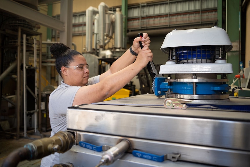 A woman wearing safety glasses works with a piece of equipment attached to a stainless steel tank.